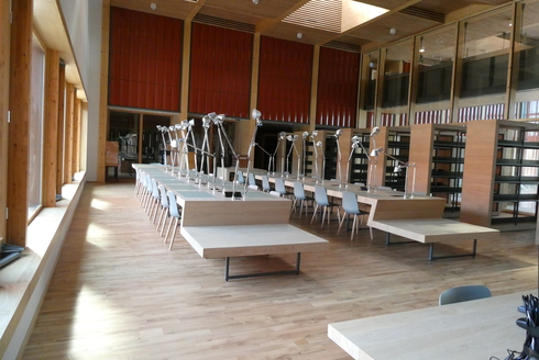 Vue sur une salle de lecture équipée de menuiserie bois et d'un plancher bois, 2 grandes rangées de bureau équipée de lampes orientables et sur la droite, des rangées vides de bibliothèques - Agrandir l'image (fenêtre modale)