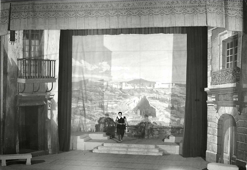Photo d'un homme en costume de théâtre se tient au milieu dans un décor qui figure une rue, des façades de maison avec balconnet de part et d'autres.