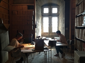 Dans une vieille salle pavée, à la lueur d'une ancienne fenêtre à meneaux, deux archivistes classent des documents. - Agrandir l'image, .JPG 2,3 MB (fenêtre modale)